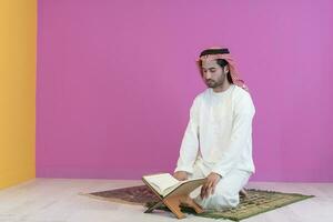 jovem árabe muçulmano homem dentro tradicional roupas lendo piedosos livro Alcorão em a Rezar tapete antes iftar jantar durante uma Ramadã festa às casa foto