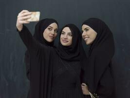 grupo do jovem muçulmano mulheres dentro elegante vestir com hijab usando Smartphone enquanto levando selfie cenário dentro frente do Preto fundo foto