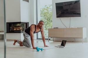 Treinamento às lar. desportivo homem fazendo Treinamento enquanto assistindo conectados tutorial em computador portátil, exercício dentro vivo sala, livre espaço foto