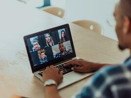 africano americano homem dentro óculos sentado às uma mesa dentro uma moderno vivo sala, usando uma computador portátil para o negócio vídeo bater papo, conversação com amigos e entretenimento foto