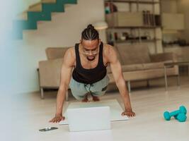 jovem africano americano homem trabalhando flexões dentro a vivo quarto enquanto assistindo conectados Treinamento em computador portátil foto