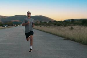 atraente em forma homem corrida velozes ao longo campo estrada às pôr do sol luz, fazendo corrida exercite-se ao ar livre foto