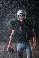 americano futebol campo solitário atleta Guerreiro em pé em uma campo detém dele capacete e pronto para jogar. jogador preparando para correr, ataque e Ponto aterragem. chuvoso noite com dramático névoa, azul luz foto