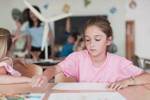 pequeno meninas sentado dentro elementar escola desenhando em papel com seus amigos enquanto sentado dentro uma moderno Sala de aula foto