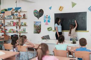 elementar escola. a fêmea professor ajudando a criança aluna enquanto escrevendo a responda em a quadro-negro. foto