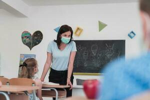 multirracial grupo do crianças vestindo face máscaras trabalhando às aula, escrevendo e ouvindo explicações do professor dentro Sala de aula foto