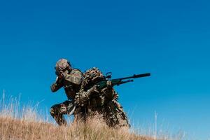 uma Franco atirador equipe pelotão do soldados é indo disfarçado. Franco atirador assistente e equipe líder caminhando e visando dentro natureza com amarelo Relva e azul céu. tático camuflar uniforme. foto