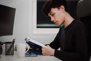 jovem bonito asiático lendo livro na mesa de trabalho tarde da noite foto