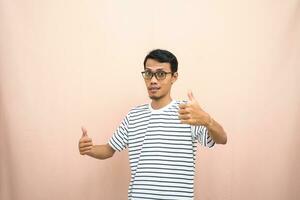 ásia homem dentro óculos vestindo casual listrado camisa, posando dando polegares acima gesto ou OK este concordar. isolado bege fundo. foto