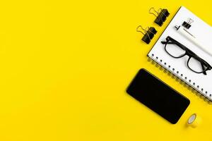 moderno escritório escrivaninha plano deitar com Smartphone tela brincar, espiral papel bloco de anotações e lendo óculos em amarelo fundo. topo visualizar. cópia de espaço. foto