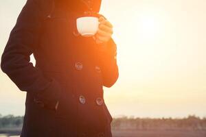mãos de mulher segurando uma xícara de café, casaco preto pela manhã foto