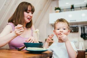alegre criança comendo massa dentro a cozinha com mãe foto