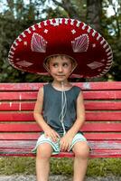 pequeno fofa Garoto vestindo uma sombrero é sentado em uma vermelho Banco ao ar livre foto