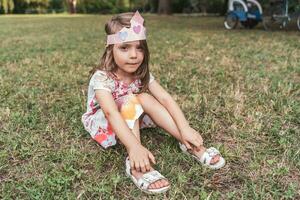 retrato do fofa menina com uma papel coroa sentado dentro uma público parque foto