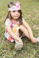retrato do fofa menina com uma papel coroa sentado dentro uma público parque foto