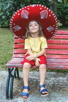pequeno fofa menina vestindo uma grande sombrero é sentado em uma vermelho Banco dentro uma público parque foto