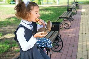 menina com mochila comendo sanduíche embalado dentro uma sanduíche caixa perto escola. uma rápido lanche com uma pão, pouco saudável comida, almoço a partir de escola. costas para escola. Educação, primário escola Aulas, setembro 1 foto