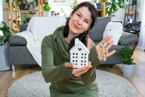 uma feliz mulher dentro dela casa detém dentro dela mãos uma miniatura figura do uma casa e chave dentro a interior. Sonhe casa projeto, real Estado comprar, seguro, hipoteca, aluguel, reserva foto