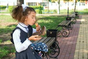 menina com mochila comendo sanduíche embalado dentro uma sanduíche caixa perto escola. uma rápido lanche com uma pão, pouco saudável comida, almoço a partir de escola. costas para escola. Educação, primário escola Aulas, setembro 1 foto