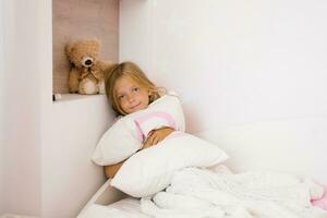 feliz fofa menina criança sentado em a cama abraçando uma travesseiro foto