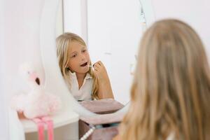 bonita criança menina tintas dela lábios com brilhar ou bálsamo dentro frente do uma espelho dentro dela quarto foto