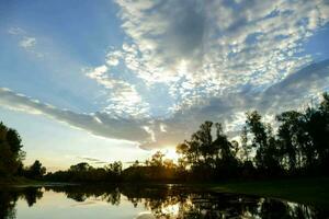 a Sol é configuração sobre uma lago com árvores e nuvens foto