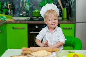 feliz pequeno Garoto preparando massa dentro cozinha às mesa. lá estão massa produtos em mesa, vestido Como chefe de cozinha foto