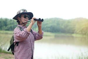 ásia Senior mulher vai viajando sozinho, usar binocular para pesquisa natureza às a lago. conceito, lazer atividade. Senior estilo de vida. gastar Tempo para viagem depois de aposentado. foto