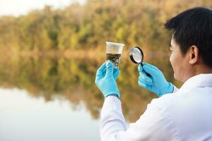 homem asiático ecologista detém lupa para inspecionar a água em vidro transparente do lago. conceito, explorar, analisar a qualidade da água e a criatura da fonte natural. pesquisa de campo em ecologia. foto