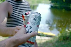 jovem homem levantar Budweiser broto Cerveja pode com masculino amigo em borrado rio com caiaque e árvores foto