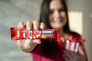 Carcóvia, Ucrânia - Outubro 21, 2019 uma jovem caucasiano morena menina mostra kit Kat chocolate Barra dentro vermelho invólucro dentro luz sala. kit Kat chocolate bens fabricado de aninhar foto