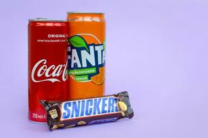 risadinhas chocolate Barra dentro Castanho invólucro mentiras com vermelho coca Cola e laranja fanta lata pode em lilás pastel fundo. famoso beber e chocolate produtos foto