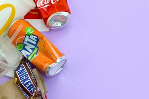 risadinhas chocolate Barra invólucro mentiras com esvaziar vermelho coca Cola e laranja fanta lata latas Próximo para mcdonalds papel bolsas e copos em lilás pastel fundo. famoso beber e chocolate produtos Lixo foto