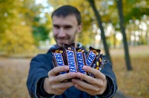 Carcóvia, Ucrânia - Outubro 8, 2019 uma jovem caucasiano barbudo homem mostra muitos risadinhas chocolate bares dentro Castanho invólucro dentro outono parque. risadinhas chocolate fabricado de Marte foto
