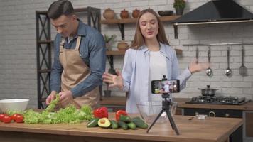 blogging online, celular produz vídeo para assinantes como casal de blogueiros cozinham preparando refeições saudáveis com vegetais frescos e verduras fechadas na mesa da cozinha