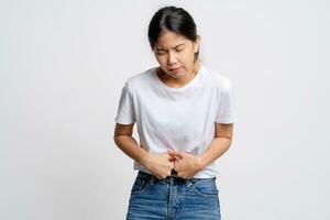 ásia mulher dentro branco camiseta tendo estômago dor em pé isolado em branco fundo. foto