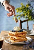 waffles coberto com amoras, gelo creme e querida em pé em uma rústico Castanho mesa. azul fundo. mão garoa mel. foto