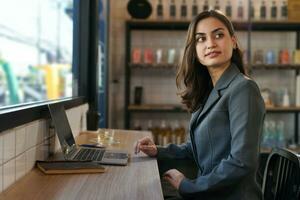 atraente o negócio mulher dentro terno sentado às contador bar, jovem moderno fêmea controlo remoto trabalhar, uma digital nômade trabalhando sobre computador portátil Internet conectados dentro cafeteria dentro de casa foto