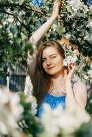 lindo jovem menina dentro uma azul vestir dentro uma florescendo maçã Pomar. florescendo maçã árvores com branco flores foto