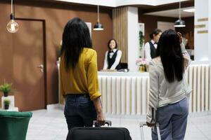 fêmea turistas A chegar às destino carregando bagagens para frente escrivaninha do moderno recorrer. turistas iniciando período de férias dentro extravagante hotel lobby pronto para faço reserva foto