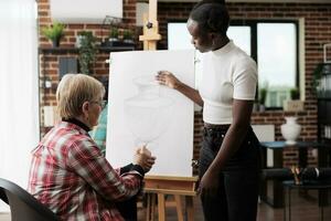 Senior mulher noivando dentro social Atividades, levando desenhando lição. africano americano jovem mulher arte instrutor ensino Mais velho adultos para desenhar dentro comunidade Centro em pé perto cavalete ajudando aluna foto