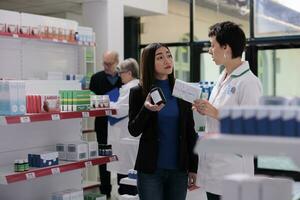 farmacêutico assistente e jovem ásia mulher discutindo Vitamina complexo componentes dentro Drogaria. farmacia empregado dando assistência para cliente dentro escolhendo multivitamínico cápsulas foto