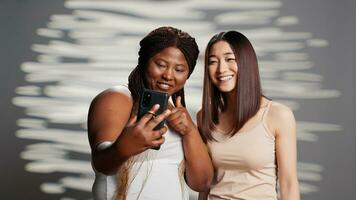 interracial alegre senhoras levando fotos com telefone, fêmea modelos promovendo diferente tons de pele e corpo tipos. confiante natural mulheres rindo com As fotos para cuidados com a pele campanha.