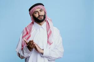 árabe homem vestido dentro tradicional islâmico roupas fazer aplausos, parabenizando e olhando às Câmera com confiante expressão. muçulmano pessoa vestindo Thobe aplaudindo estúdio retrato foto