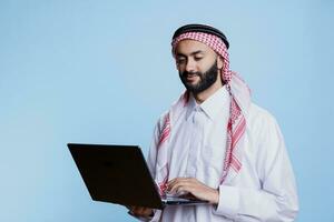 muçulmano homem vestindo tradicional Thobe e ghutra lenço de cabeça segurando computador portátil e Broswing social meios de comunicação. árabe pessoa vestido dentro islâmico roupas usando portátil computador e verificação Internet página foto