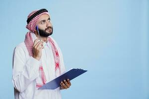 pensativo homem vestindo tradicional muçulmano roupas pensando enquanto levando notas dentro prancheta. árabe contemplando, olhando acima com pensativo expressão enquanto segurando caneta e lista de controle foto