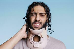 árabe homem vestindo cervical colarinho e fazendo careta dentro dor enquanto tocante pescoço. ferido jovem pessoa com doloroso facial expressão vestindo braçadeira, recuperando a partir de espinhal trauma foto