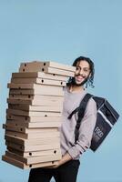 despreocupado árabe entregador carregando grande pizza caixas pilha e olhando às Câmera com alegre expressão. sorridente restaurante Entrega serviço correio em pé enorme pilha do velozes Comida pacotes foto