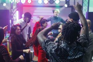 diverso pessoas dançando e levantando mãos às discoteca com viver música dentro Boate. jovem clubbers festa, comovente para eletrônico música ritmo e pulando em clube pista de dança foto