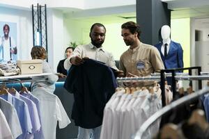 africano americano homem mostrando vestuário para roupas loja empregado e Perguntando para moda conselho. varejo mercado boutique assistente e cliente escolhendo na moda camisa juntos foto